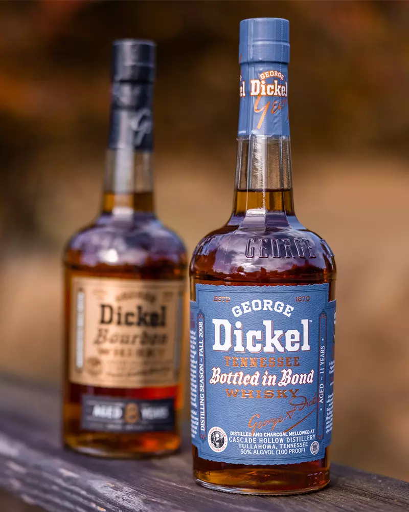 Two bottles of George Dickel whiskey are displayed on a wooden surface, with one labeled Bottled in Bond in front and a bourbon bottle slightly blurred in the background.