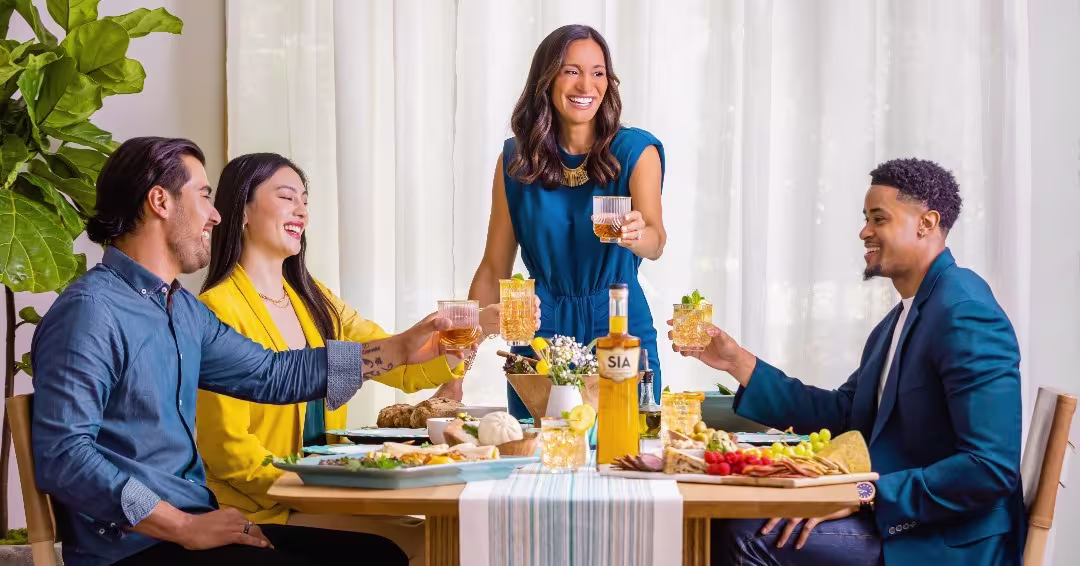 Four people sit around a table sharing drinks and food, smiling and toasting together. A bottle of SIA whiskey is on the table.