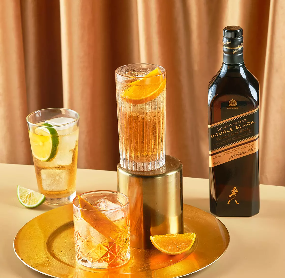 A bottle of Johnnie Walker Double Black whisky is shown with three whisky cocktails garnished with citrus slices, placed on a gold tray and table.