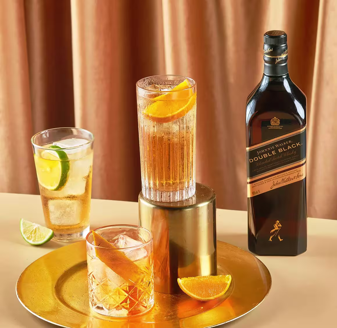 A bottle of Johnnie Walker Double Black whisky is shown with three whisky cocktails garnished with citrus slices, placed on a gold tray and table.
