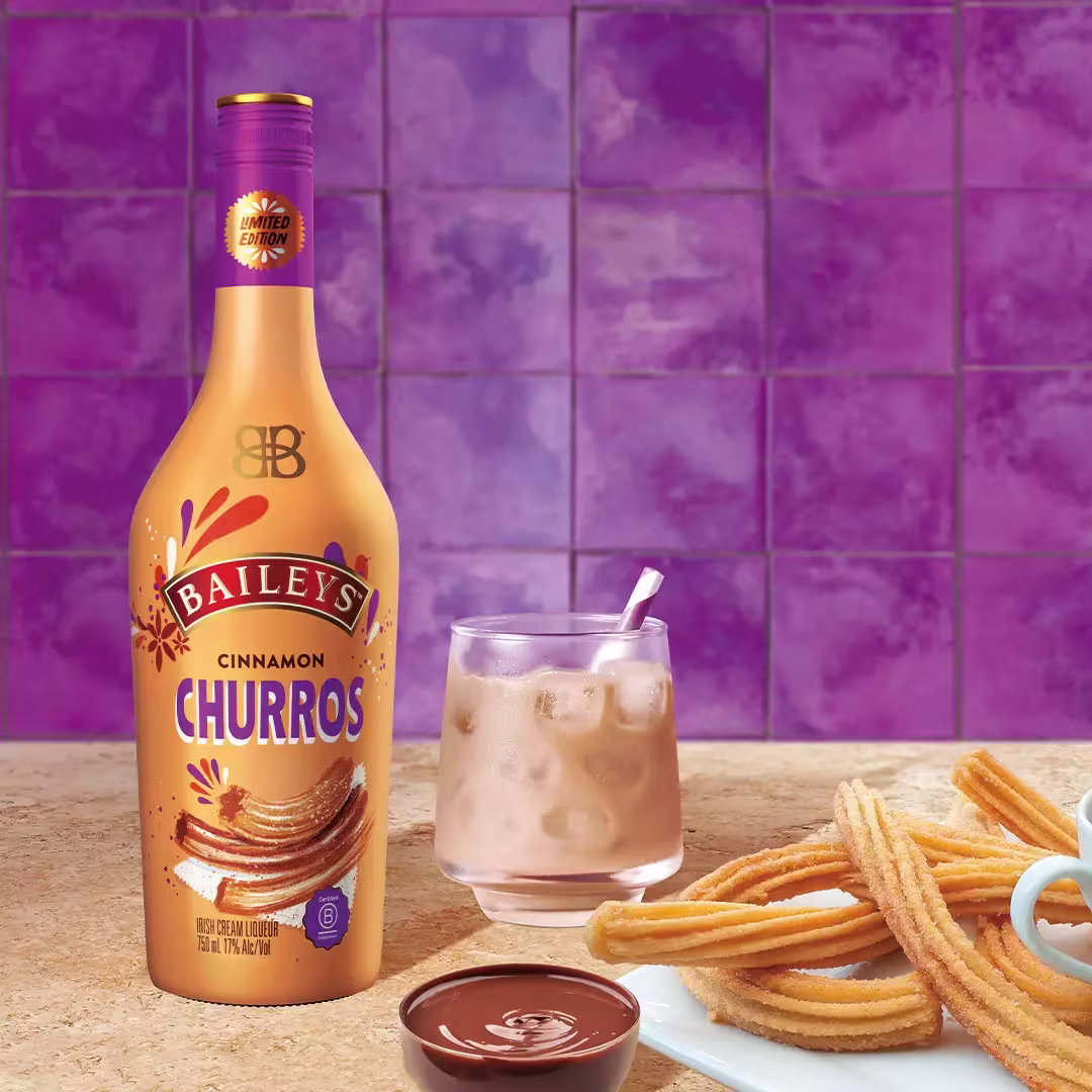 A bottle of Baileys Cinnamon Churros liqueur stands next to a glass of iced drink, a plate of churros, and a small bowl of chocolate sauce on a countertop with a purple tiled background.