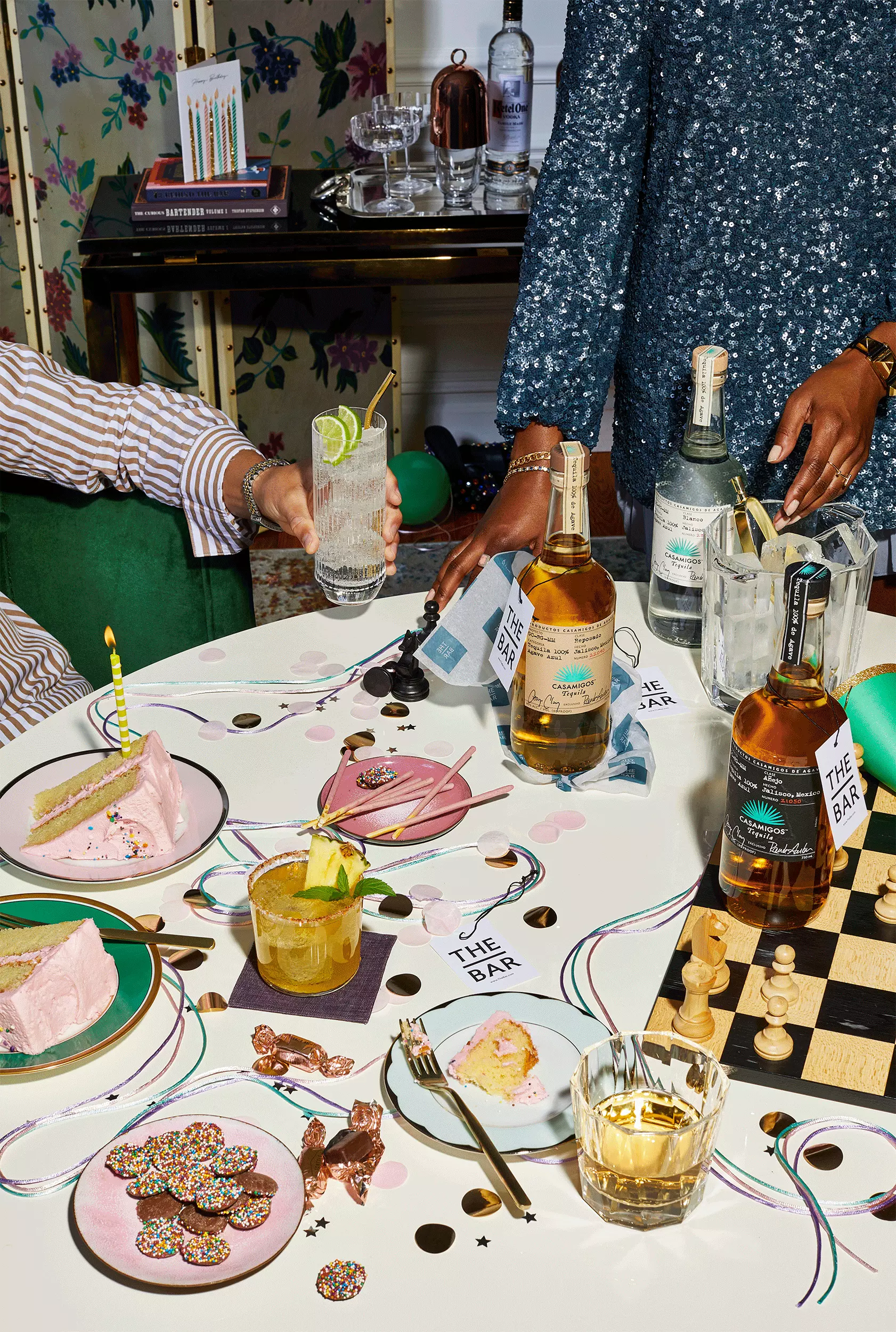 A table set for a celebration with drinks, cake, confetti, party favors, and a chessboard. Two people are standing nearby, one holding a cocktail. Bottles of alcohol are on the table.