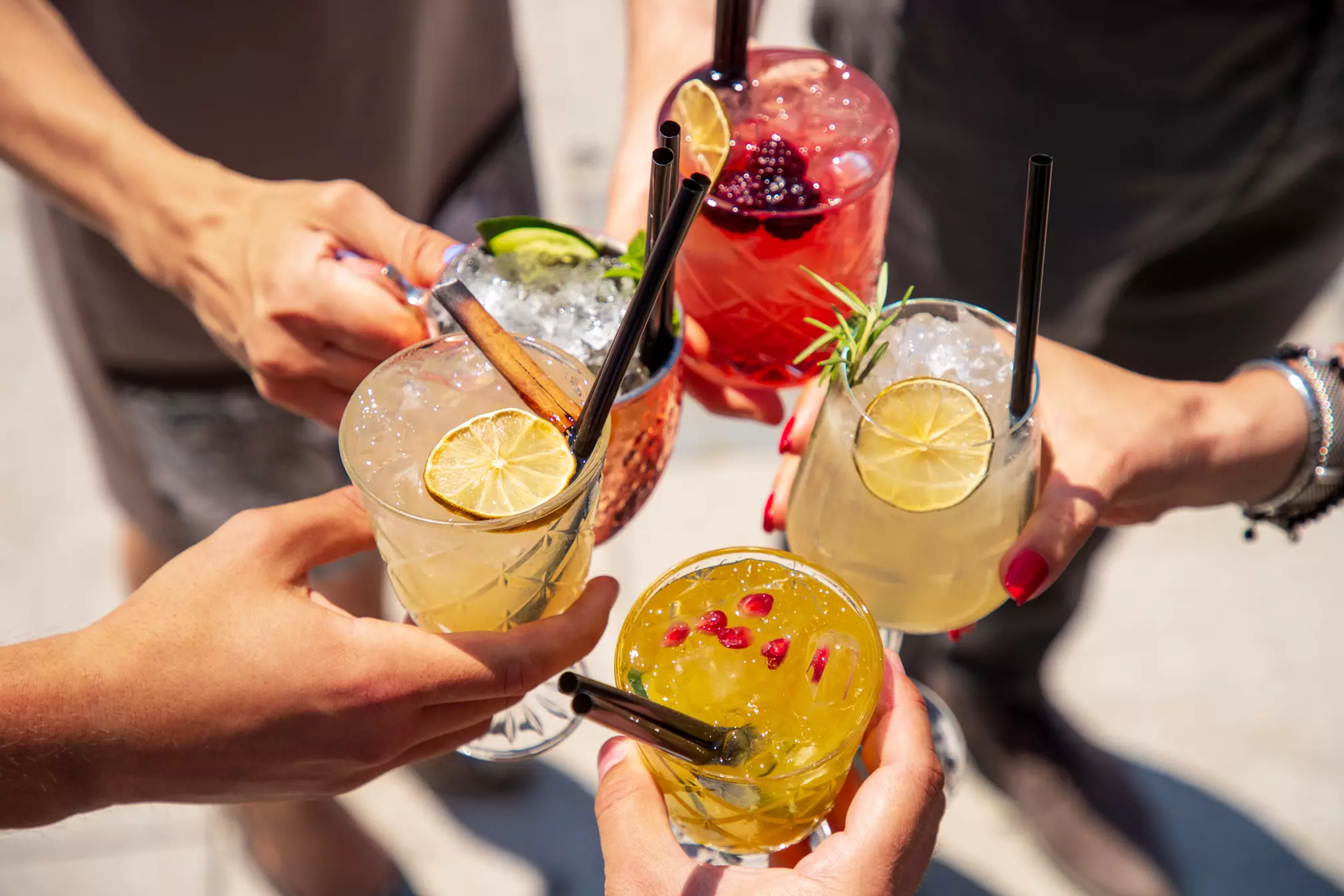 Five people hold up assorted cocktails with ice, garnishes of citrus, berries, herbs, and pomegranate seeds, viewed from above in an outdoor setting.