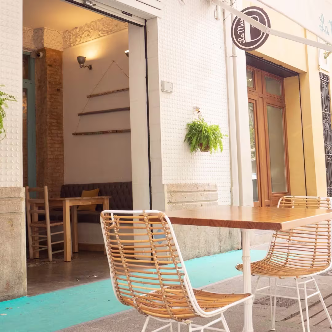 La Más Bonita luce una fachada mediterránea con detalles en azul pastel, sillas de mimbre y una atmósfera acogedora perfecta para cualquier hora del día.