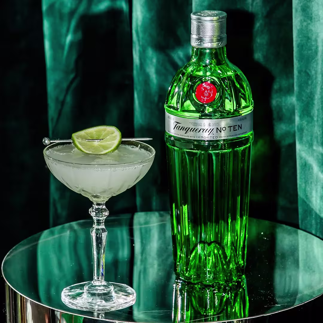 A bottle of Tanqueray No. Ten gin stands next to a cocktail glass with a lime wedge on a reflective round surface, against a green velvet background.