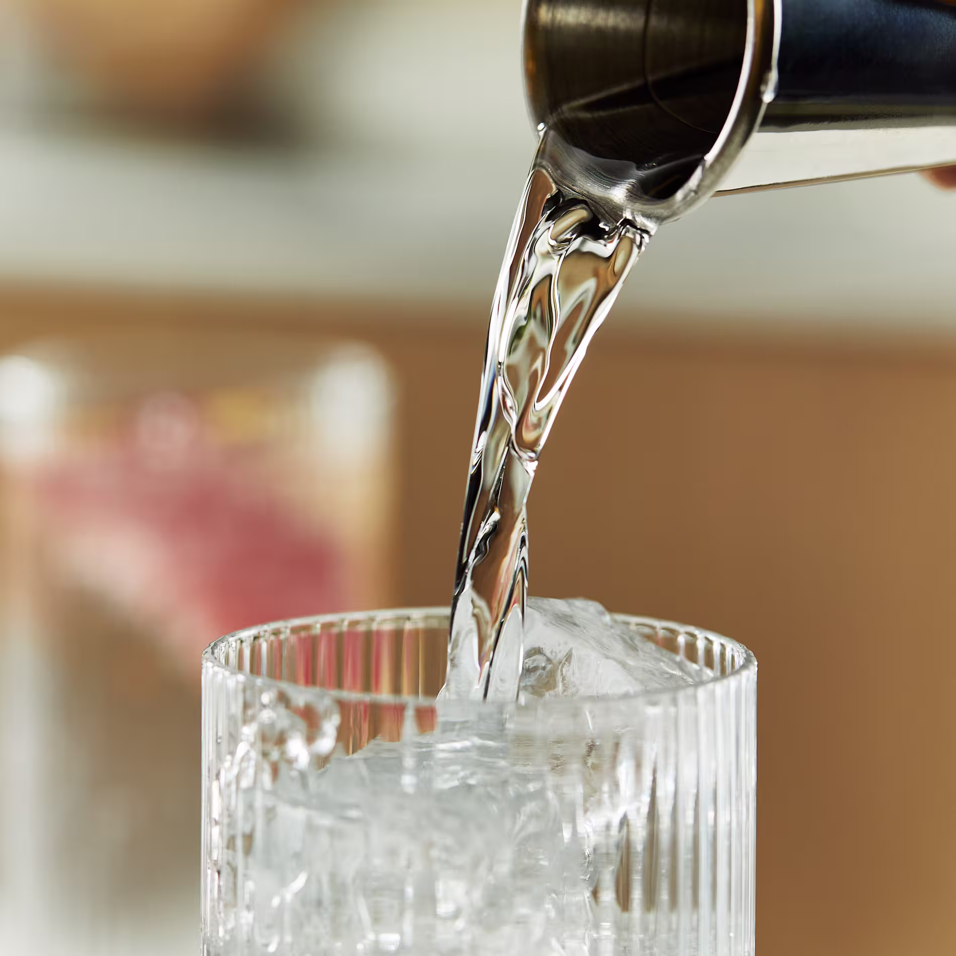A clear liquid is poured from a silver jigger into a highball glass filled with ice