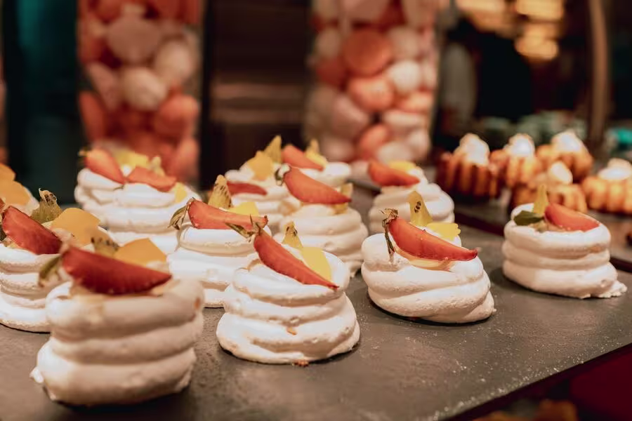 Small meringue desserts topped with sliced fruit are arranged on a tray, with other assorted pastries visible in the background.