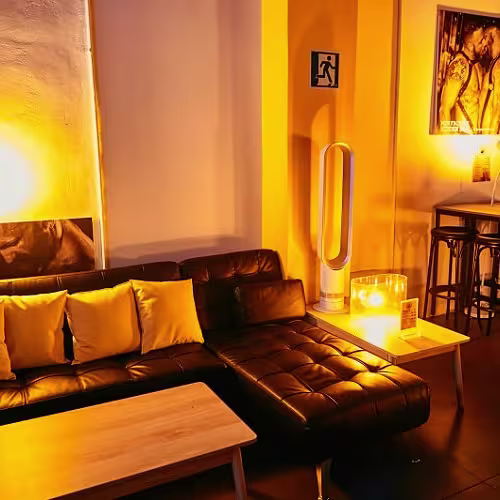 Sala con sofá negro, luz cálida, mesa de madera y póster en pared. Ambiente acogedor con decoración íntima y mesas altas al fondo.