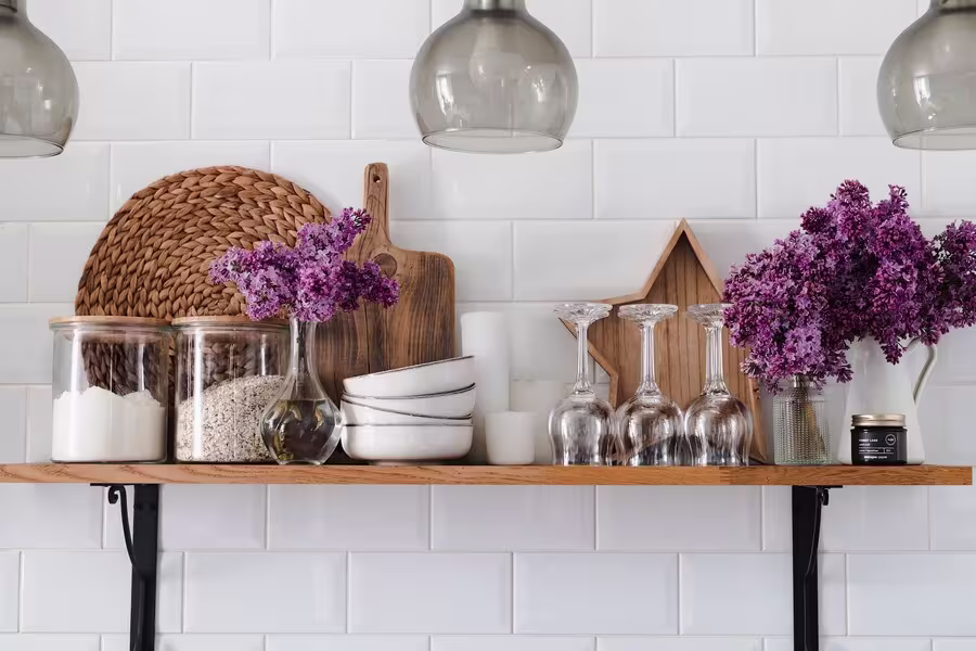 A wooden kitchen shelf with glass jars, white bowls, wine glasses, cutting boards, a decorative star, and vases of purple flowers against a white tiled wall.