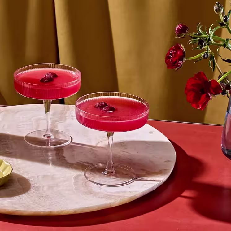 Two coupe glasses filled with bright red cocktails, garnished with berries, are placed on a round marble tray; a vase with red flowers is in the background.