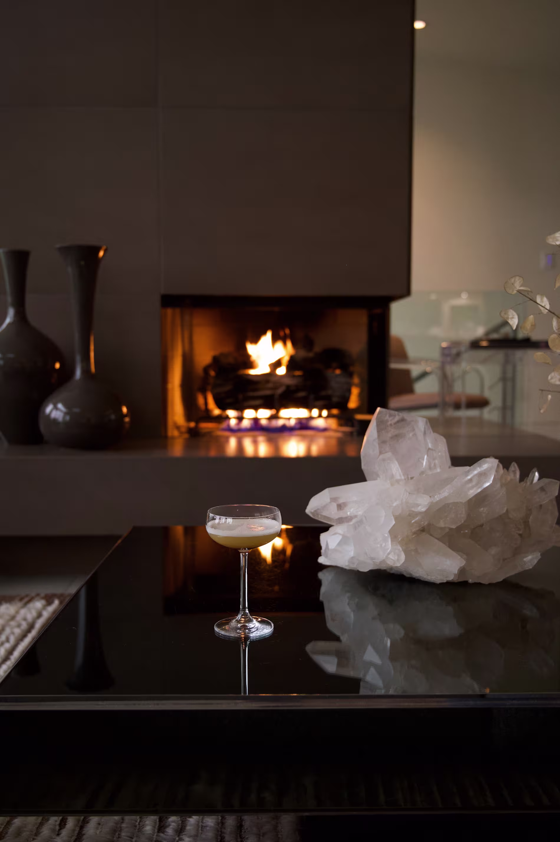 A cocktail glass sits on a black glass coffee table next to a large quartz crystal, with a lit fireplace and decorative vases in the background.