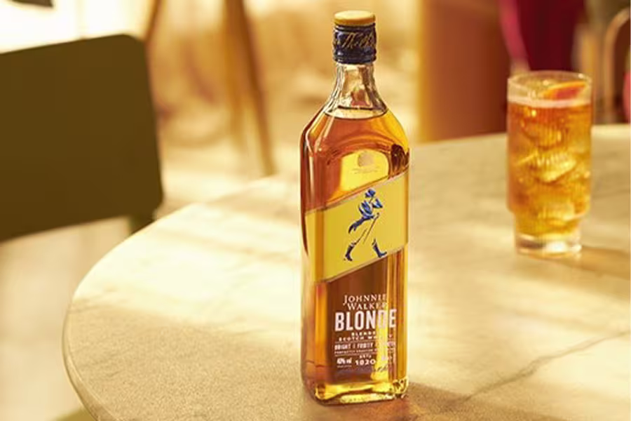 A bottle of Johnnie Walker Blonde whisky stands on a table next to a glass filled with a light brown drink and ice.