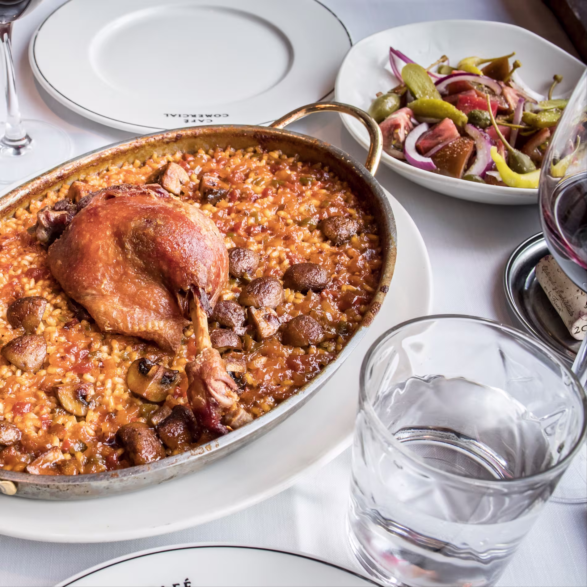 La imagen muestra una mesa con un plato del Café Comercial En el centro hay una gran paellera que contiene paella, caracterizada por su arroz amarillo-naranja y una variedad de ingredientes como verduras y carne. Encima de la paella hay un ave cocida.