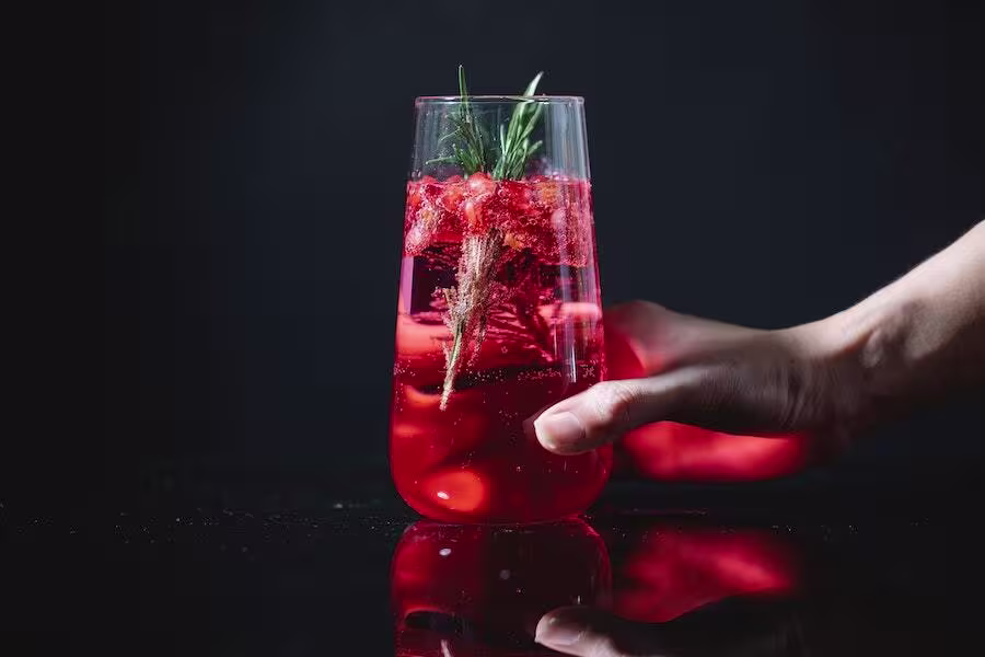 A hand holds a tall glass filled with red iced beverage, garnished with a sprig of rosemary, against a dark background.