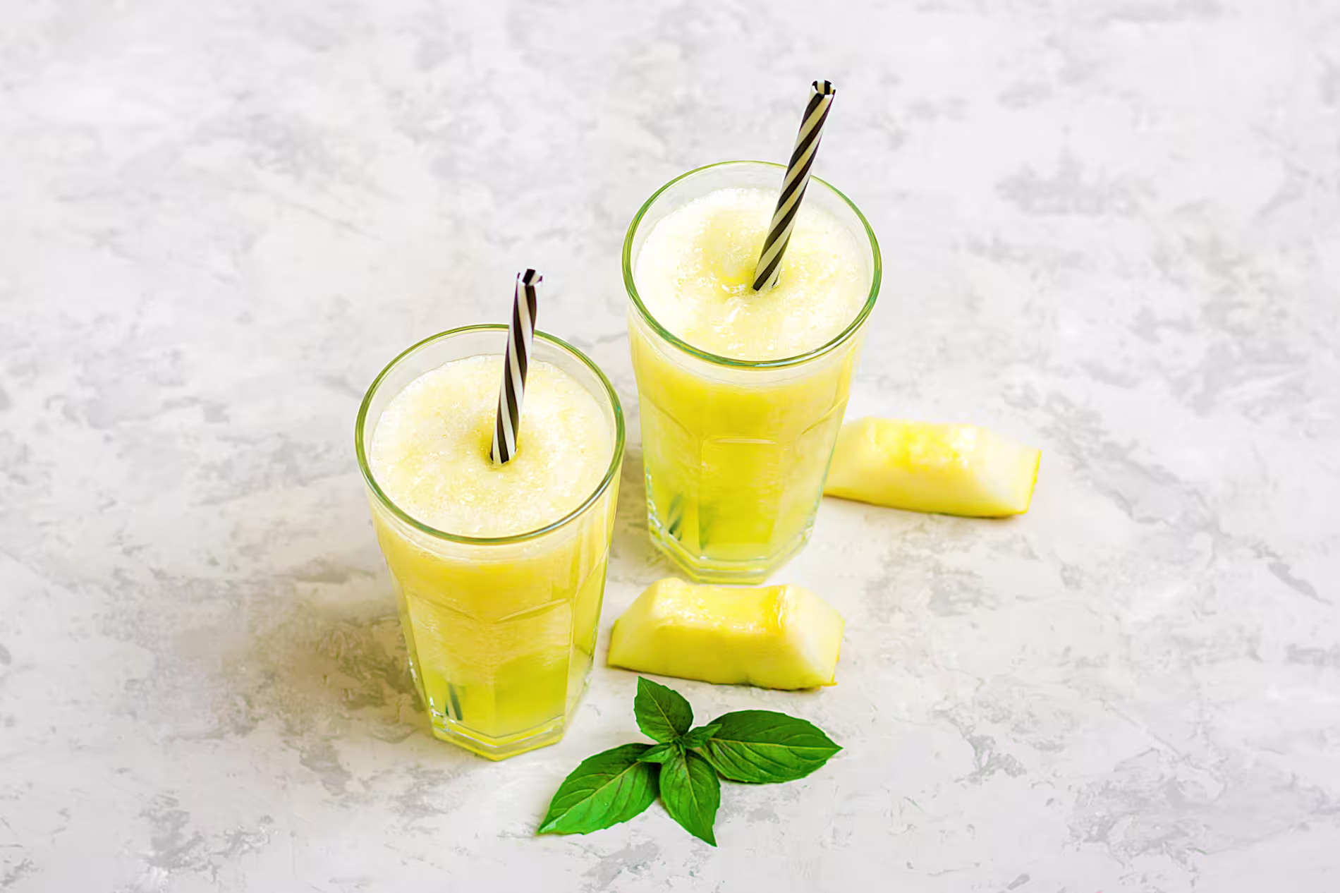 Two glasses of frozen yellow pineapple drink with black and white striped straws, garnished with pineapple slices and basil leaves on a light textured surface.