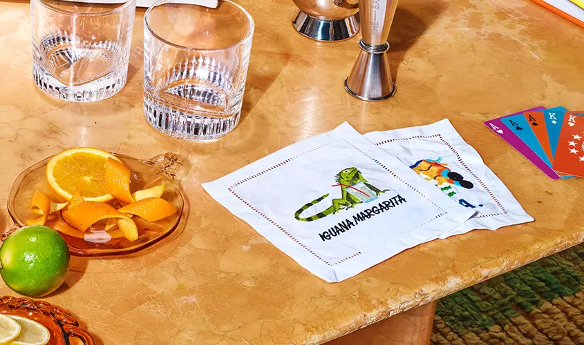 Glasses, orange slices, and a lime on a marble table next to cocktail napkins labeled Iguana Margarita and a fan of colorful playing cards.