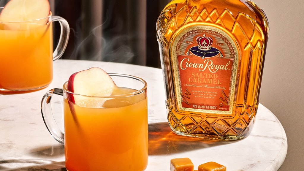 A bottle of Crown Royal Salted Caramel whisky sits on a marble table next to two glass mugs of orange drink garnished with apple slices and two caramel candies.