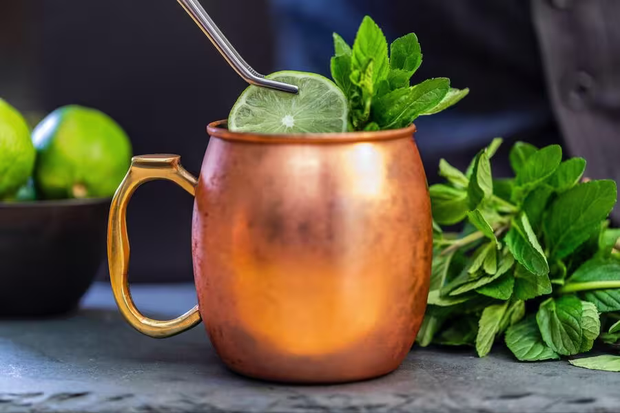 A copper mug filled with a cocktail, garnished with a lime slice and fresh mint, with a metal straw and more mint and limes nearby.