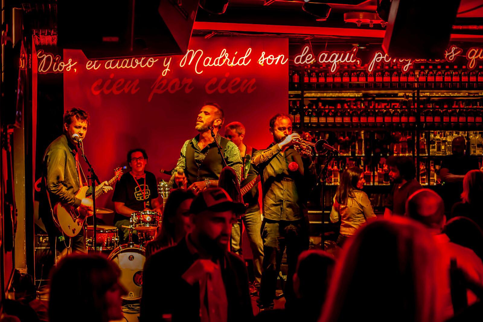Concierto íntimo en Cien por Cien Bar, con banda en vivo y luces rojas que destacan el lema: "Dios, el diablo y Madrid son de aquí.