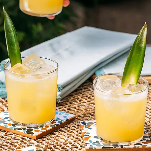 Two glasses of yellow cocktail with ice and pineapple leaf garnish, placed on colorful coasters on a woven surface, with napkins in the background.