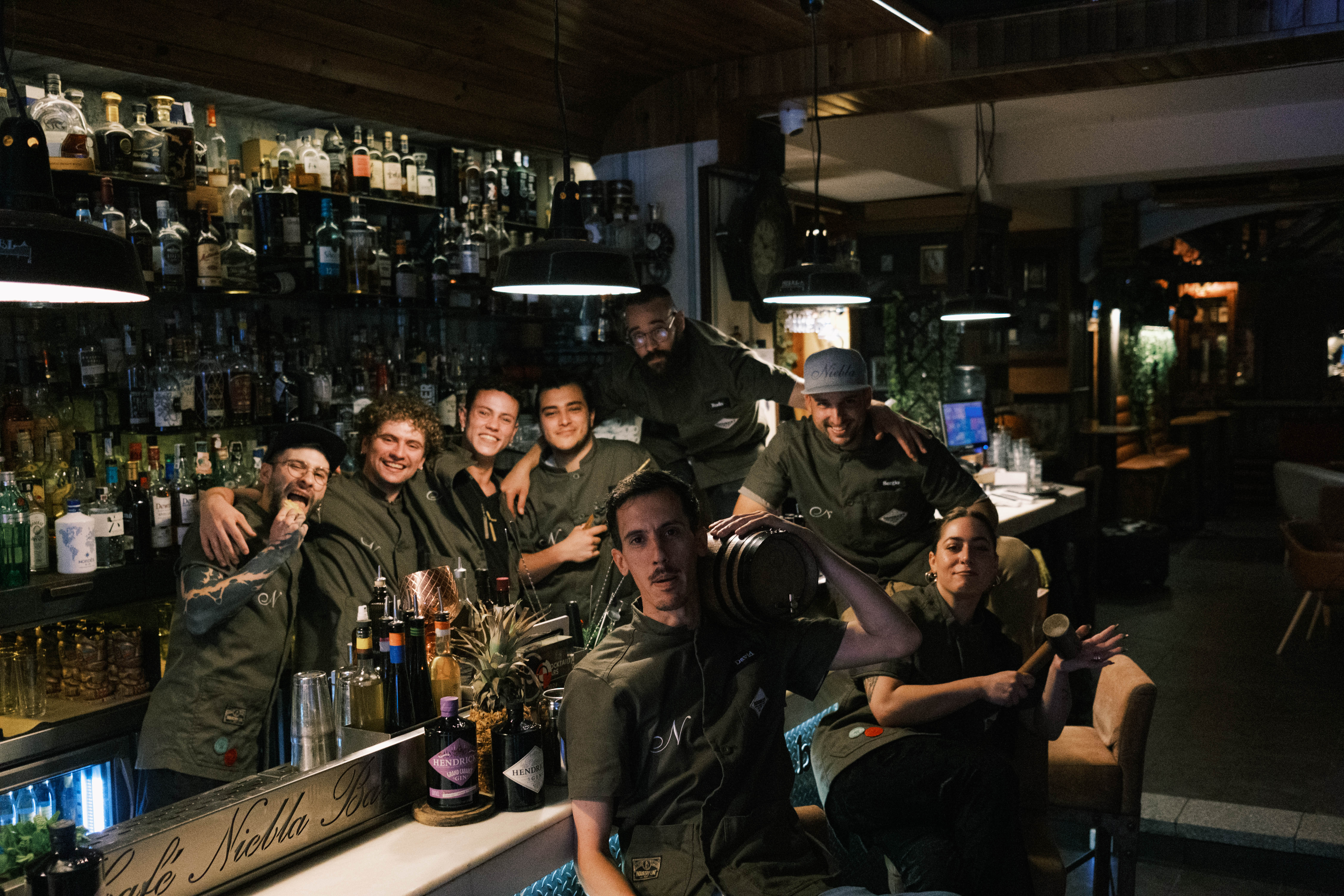 Equipo del bar Niebla posando detrás de la barra con ambiente acogedor, rodeados de botellas y detalles decorativos únicos