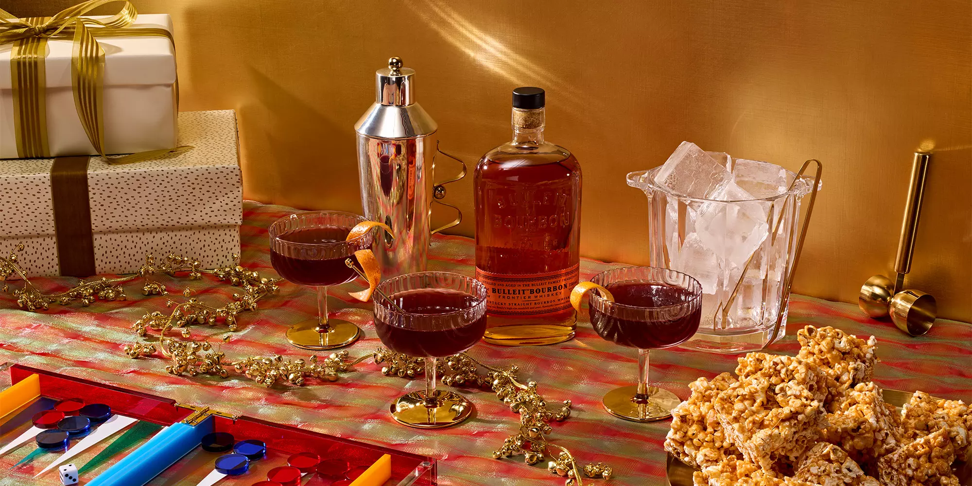 A festive table with wrapped gifts, a cocktail shaker, a bottle of Bulleit bourbon, three cocktails, an ice bucket, gold jigger, popcorn, and a backgammon game.