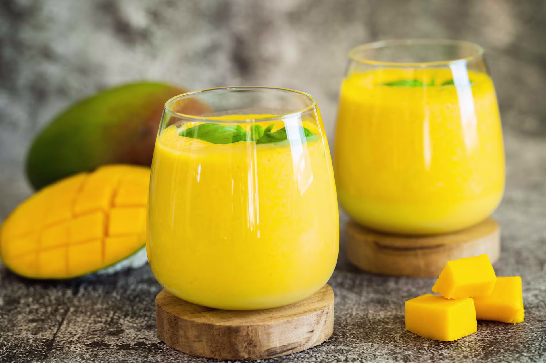Two glasses of mango smoothie on wooden coasters, with a whole mango, a cut mango, and mango cubes in the background.
