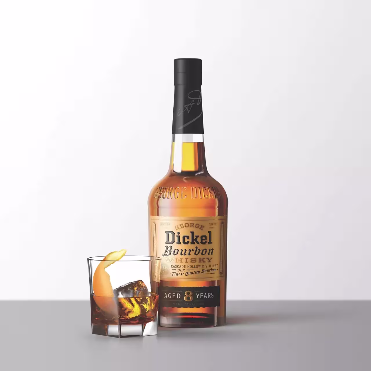 A bottle of George Dickel Bourbon Whiskey aged 8 years stands next to a glass of whiskey with ice and an orange peel garnish on a gray surface.