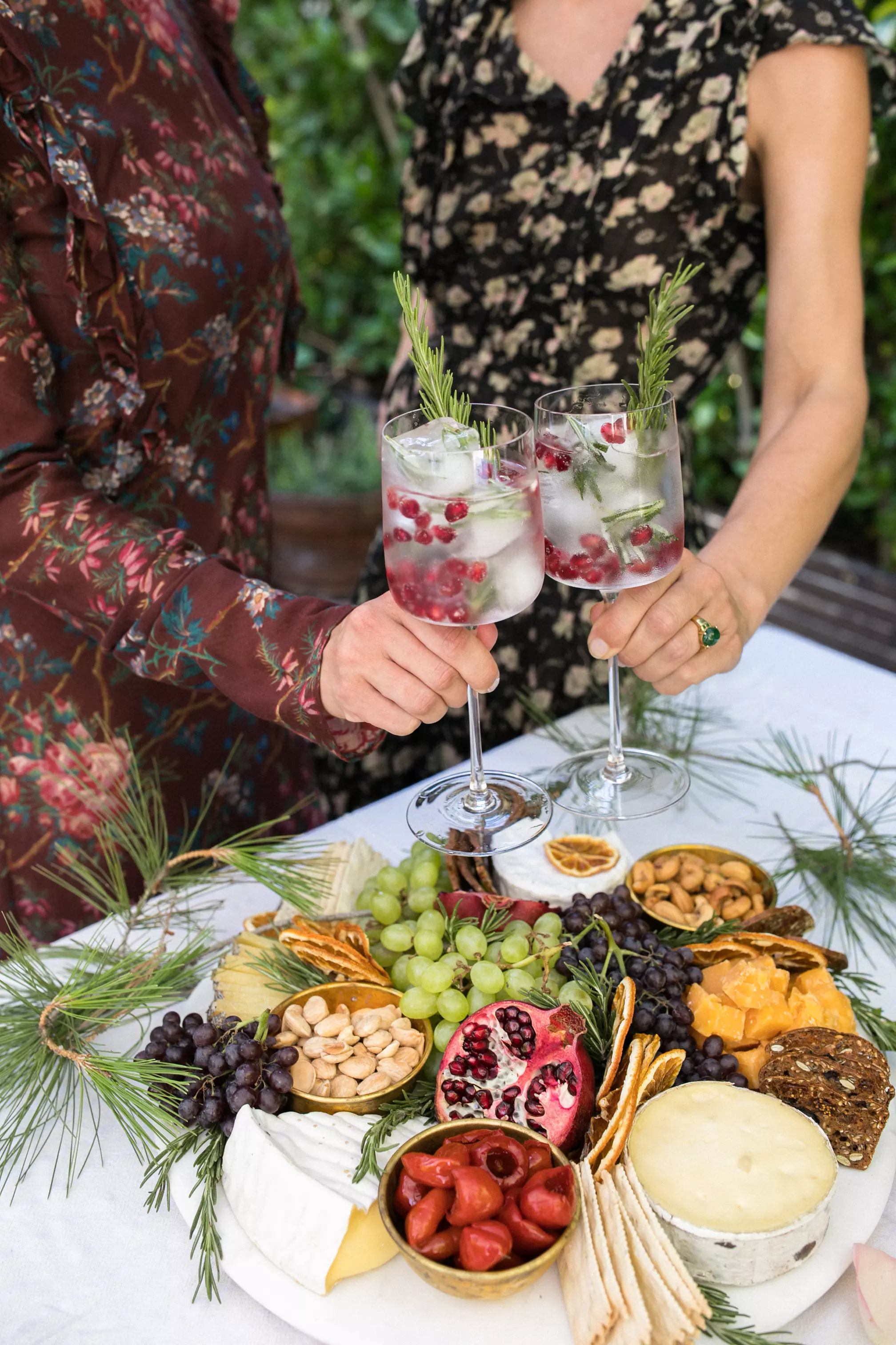 Two people clink glasses of a clear cocktail with pomegranate seeds and rosemary above a table with a charcuterie board of cheese, nuts, crackers, fruit, and pomegranate.