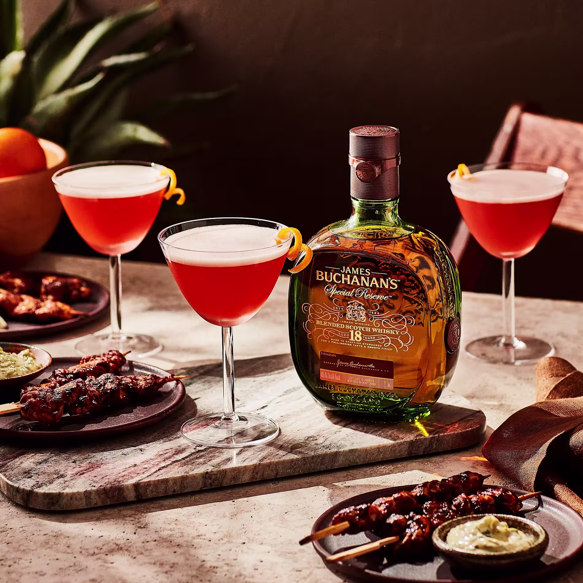 A bottle of Buchanan’s 18 Special Reserve whisky is displayed on a table with three cocktails, appetizers, and dipping sauces.