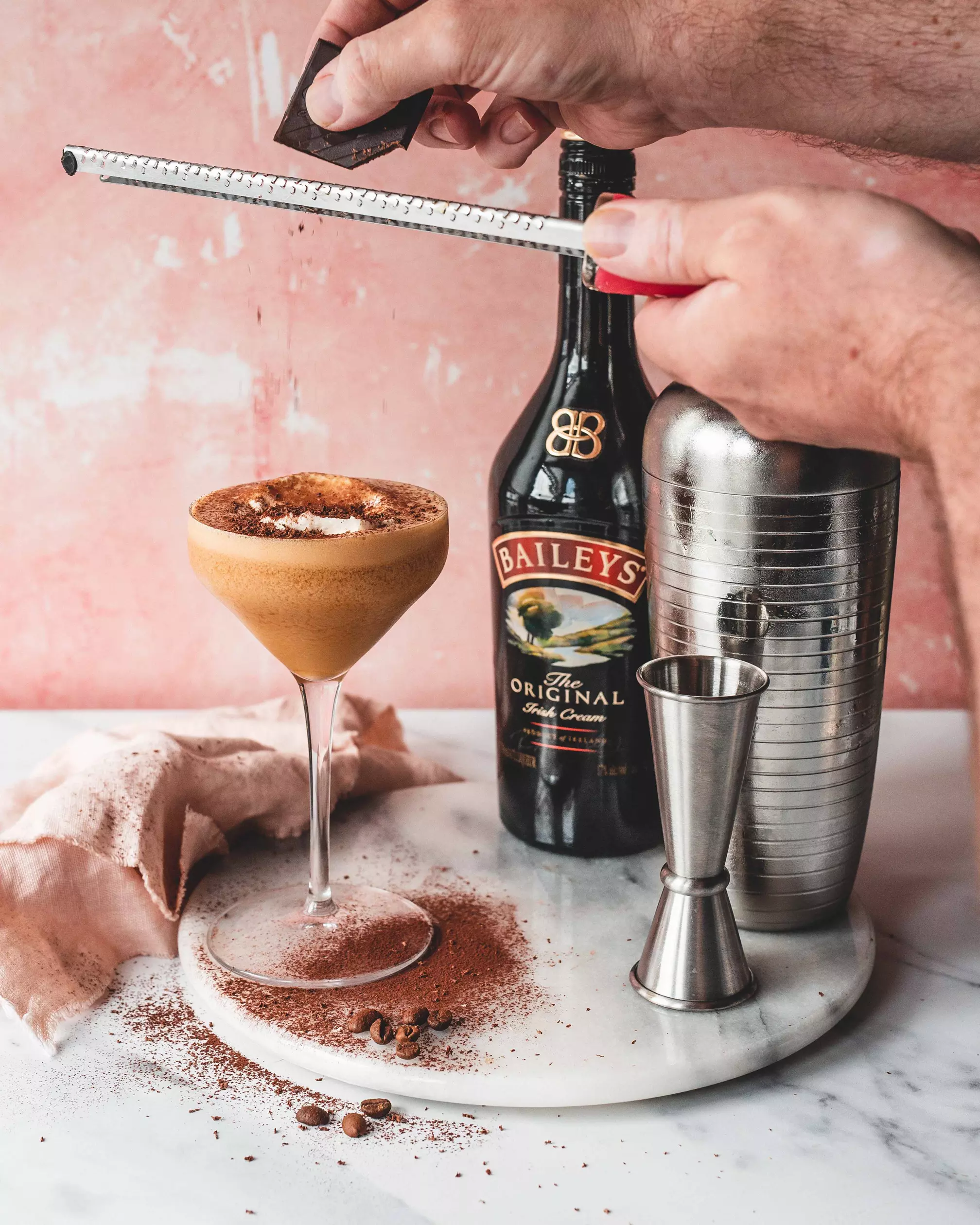 A person grates chocolate over a creamy cocktail in a coupe glass, with a Baileys bottle, cocktail shaker, and jigger on a marble surface. Cocoa powder and coffee beans are scattered.