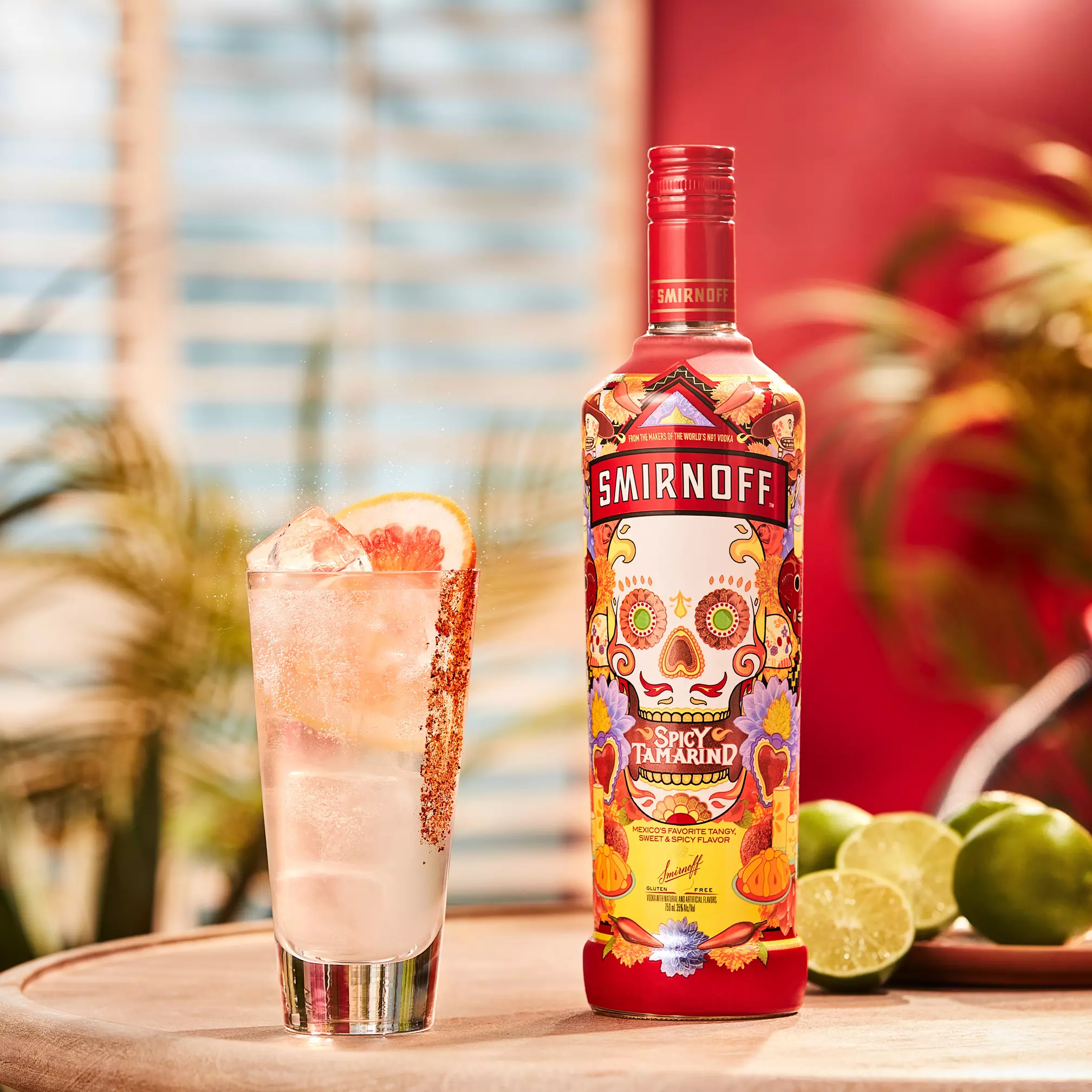 A bottle of Smirnoff Spicy Tamarind vodka stands next to a tall cocktail glass with ice, a lime wedge, and a rim dusted with spice, with limes in the background.