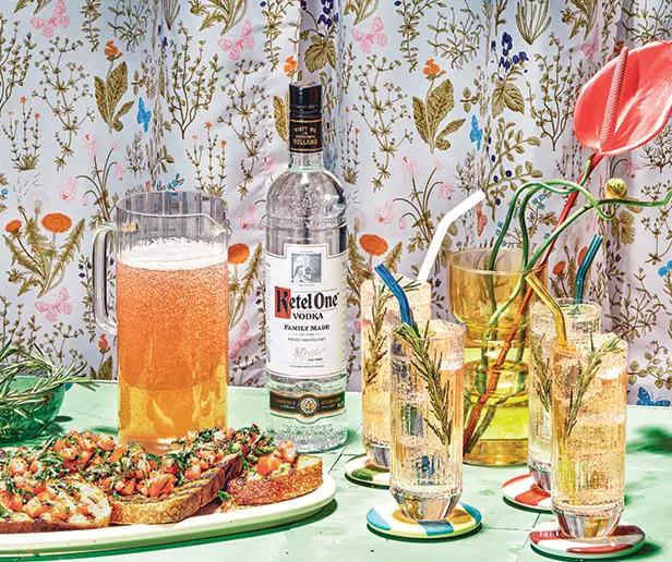A bottle of Ketel One vodka, a pitcher of a fizzy drink, three cocktails with straws, and bruschetta on a green table with a floral-patterned background.