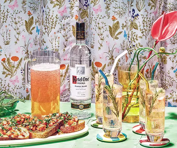 A bottle of Ketel One vodka, a pitcher of a fizzy drink, three cocktails with straws, and bruschetta on a green table with a floral-patterned background.