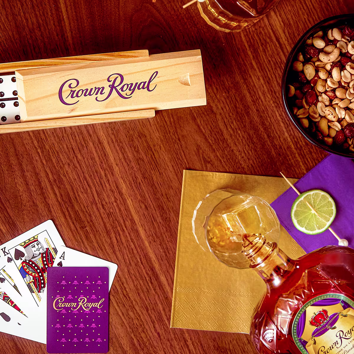 A bottle of Crown Royal, a glass with lime, playing cards, a wooden domino set, and a bowl of mixed nuts are arranged on a wooden table.