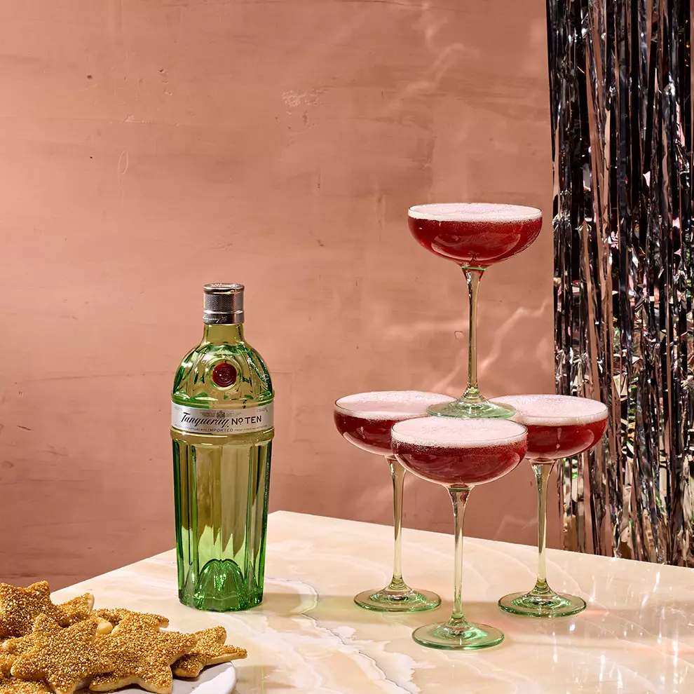 A bottle of Tanqueray No. Ten gin on a marble surface next to four coupe glasses filled with a red cocktail; gold star-shaped cookies are on a plate nearby.