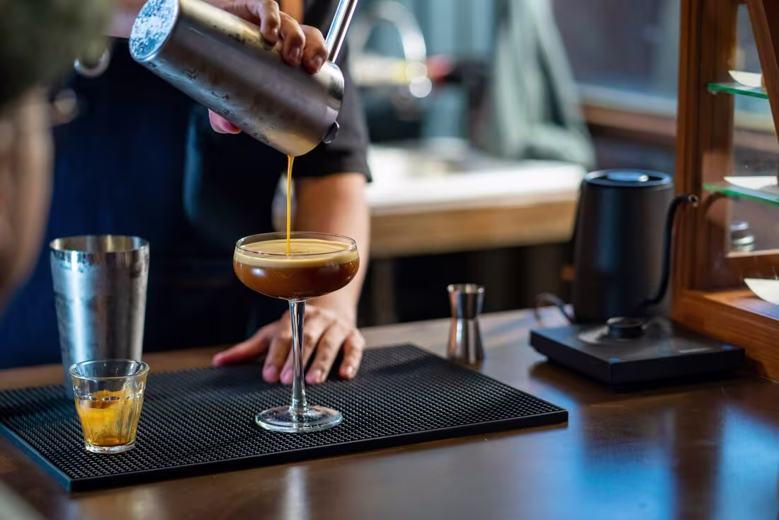 A person pours a brown cocktail from a shaker into a coupe glass on a bar counter, with various bar tools nearby.