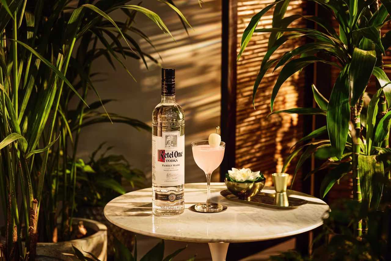 A bottle of Ketel One vodka and a pink cocktail with a garnish sit on a small round marble table, surrounded by green plants in warm lighting.