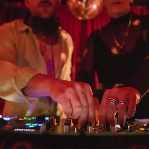 Platea: Primer plano de dos DJs manejando la consola de mezclas en un ambiente íntimo. La iluminación cálida y la bola de discoteca en el fondo crean un ambiente festivo y elegante.
