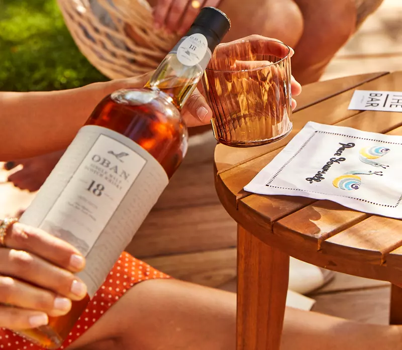 A person pours Oban 18-year-old Scotch whisky into a glass next to a wooden table with napkins and another persons hand visible.