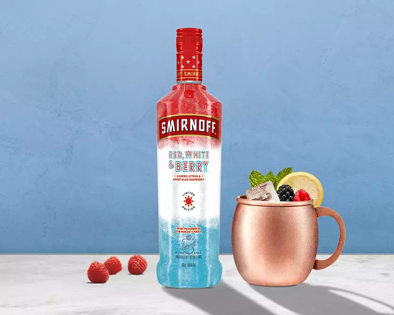 A bottle of Smirnoff Red, White & Berry vodka stands next to a copper mug filled with a mixed drink, ice, lemon slice, and berries, with three raspberries on the surface.