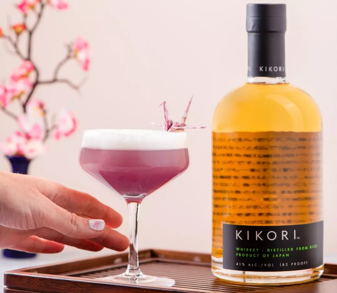 A hand holds a purple cocktail with frothy top and origami decoration, next to a bottle of Kikori whiskey on a wooden tray; a floral arrangement is blurred in the background.
