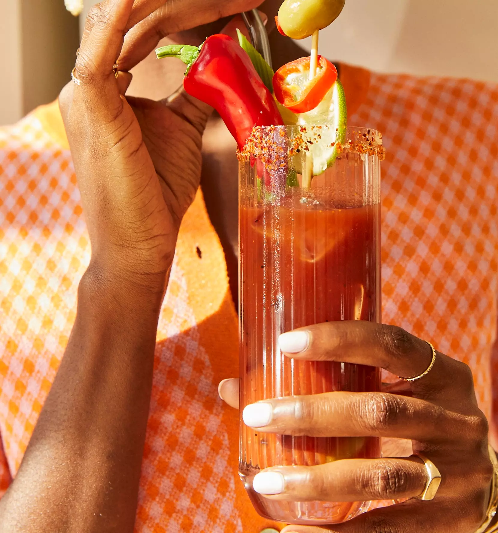 A person holds a Bloody Mary cocktail garnished with celery, peppers, cucumber, and olives. The glass rim is coated with spice, and the person wears an orange patterned shirt.