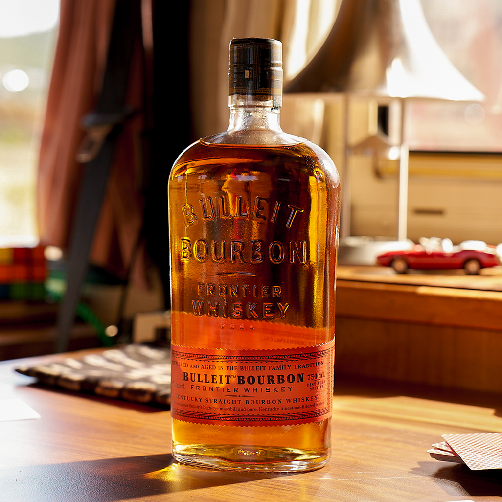 Bulleit Bourbon Frontier Whiskey bottle on a sunlit wooden table with playing cards