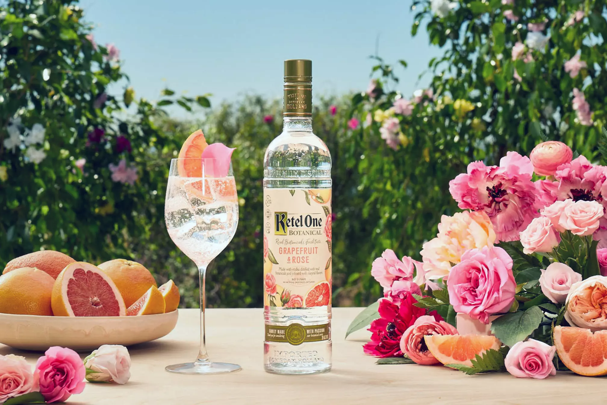 A bottle of Ketel One Botanical Grapefruit & Rose vodka stands next to a glass of cocktail, a plate of sliced grapefruit, and an arrangement of pink flowers on an outdoor table.