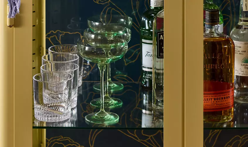 Glassware and bottles of liquor are displayed on a glass shelf inside a cabinet with a gold frame and a floral-patterned dark blue background.