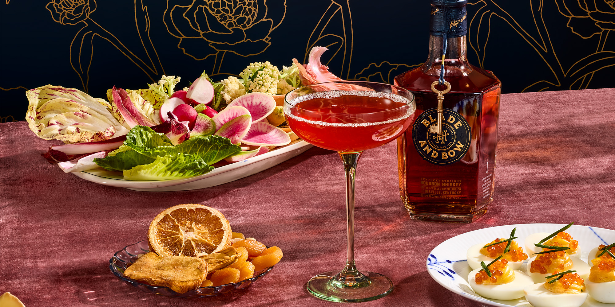A cocktail in a coupe glass sits beside a bottle of Blade and Bow bourbon, a plate of fresh vegetables, dried fruits, and deviled eggs with garnish on a pink tablecloth.