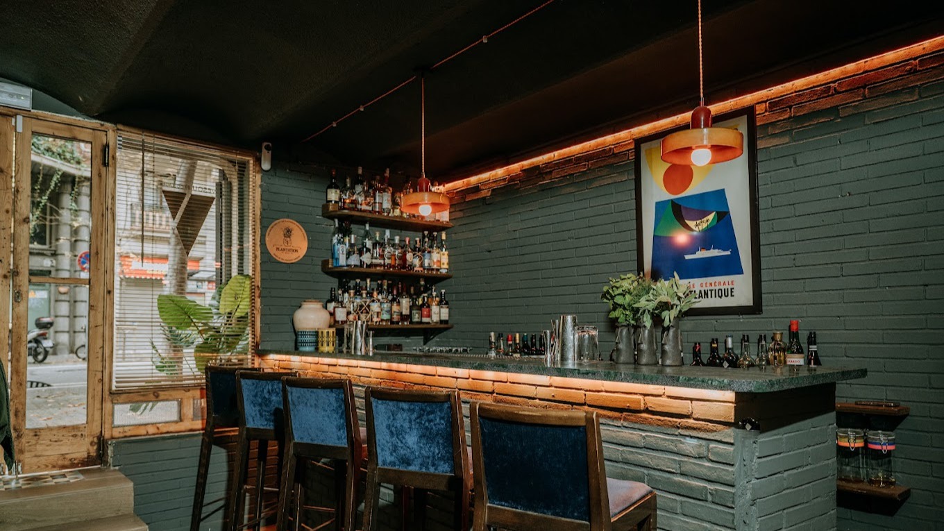 La imagen muestra el interior de un bar con un ambiente íntimo. Hay estantes con botellas y un diseño elegante, con luces cálidas que iluminan el espacio. El lugar tiene una decoración moderna y sofisticada.
