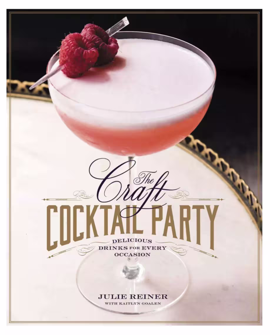 A coupe glass with a pink cocktail garnished with raspberries sits on a marble surface. The book title “The Craft Cocktail Party” by Julie Reiner is displayed over the glass.