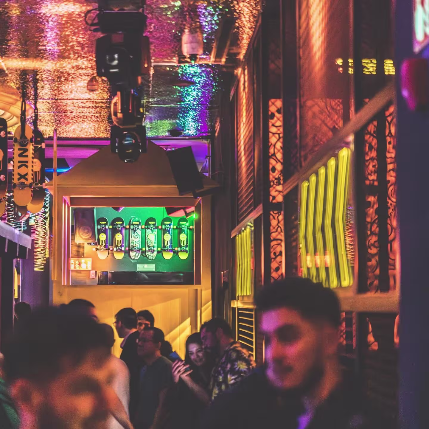 Imagen de HYP3 conambiente nocturno y animado en un club, con luces de colores que crean un efecto brillante en el techo. Varias personas socializan y se ven detalles de decoración, como monopatines en la pared.